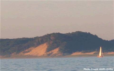 A forested sand dune at sunset sits adjacent to a lake, with a sailboat passing by.